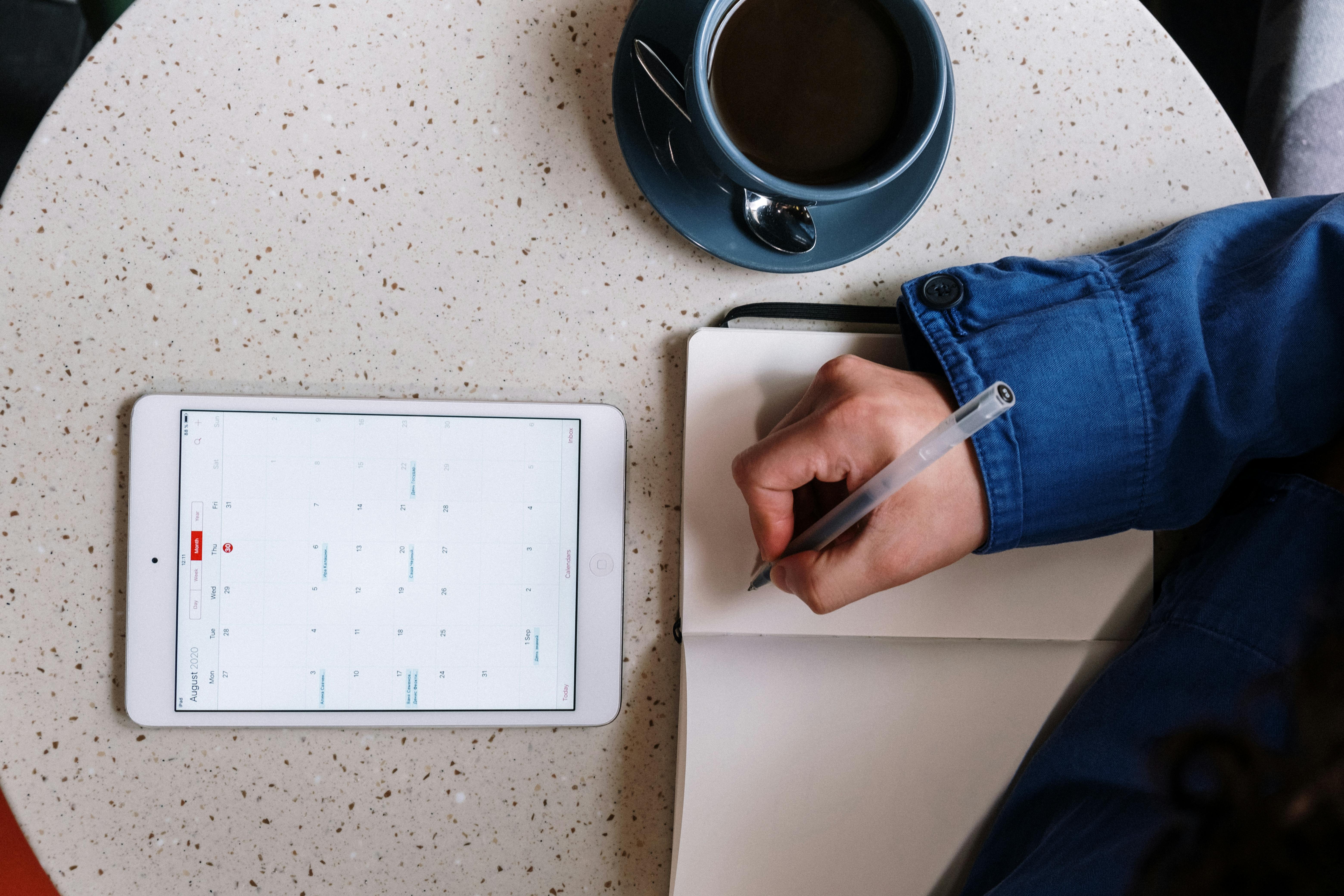 Dargestellt ist eine Person, die in einem Café mit einer Kaffeetasse Notizen aus einem digitalen Kalender macht